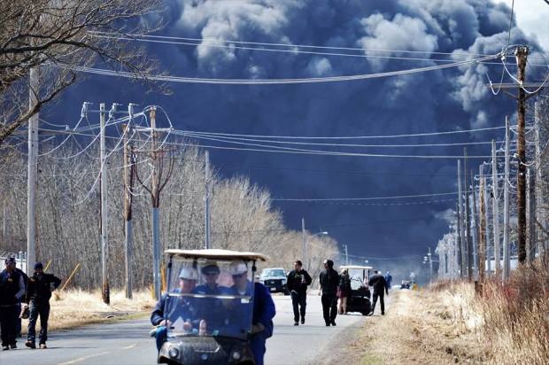 Photo shows black cloud of billowing smoke with workers walking away toward the camera, and many power lines.