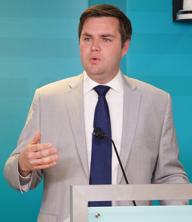 JD Vance, a white man, with a blue tie and light gray jacket, with his mouth open at a podium. 