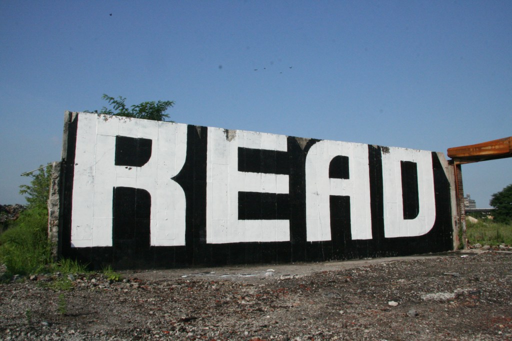 A cement wall stands alone on an abandoned lot with a blue sky. In huge letters filling the wall, graffiti text says "read."